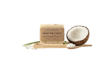 Uma barra de sabonete artesanal de óleo de coco, com rótulo impresso 'ÓLEO DE COCO', 'SABONETE ARTESANAL', '100% NATURAL • HIDRATANTE', 'FITOTERÁPICO • VEGANO'. O sabonete está sobre uma tábua de madeira, ao lado de meio coco fresco, uma colher de madeira com flocos de coco e flocos soltos com folhas verdes, contra um fundo branco limpo.