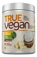 Pote de 450g do suplemento proteico True Vegan da marca True Source, sabor Chocolate Branco com Coco. O rótulo branco e laranja destaca 23g de proteína vegetal, 5,5g de BCAA e Vitamina B12, acompanhado da ilustração de um coco partido ao meio, folhas verdes e pedaços de chocolate branco.
